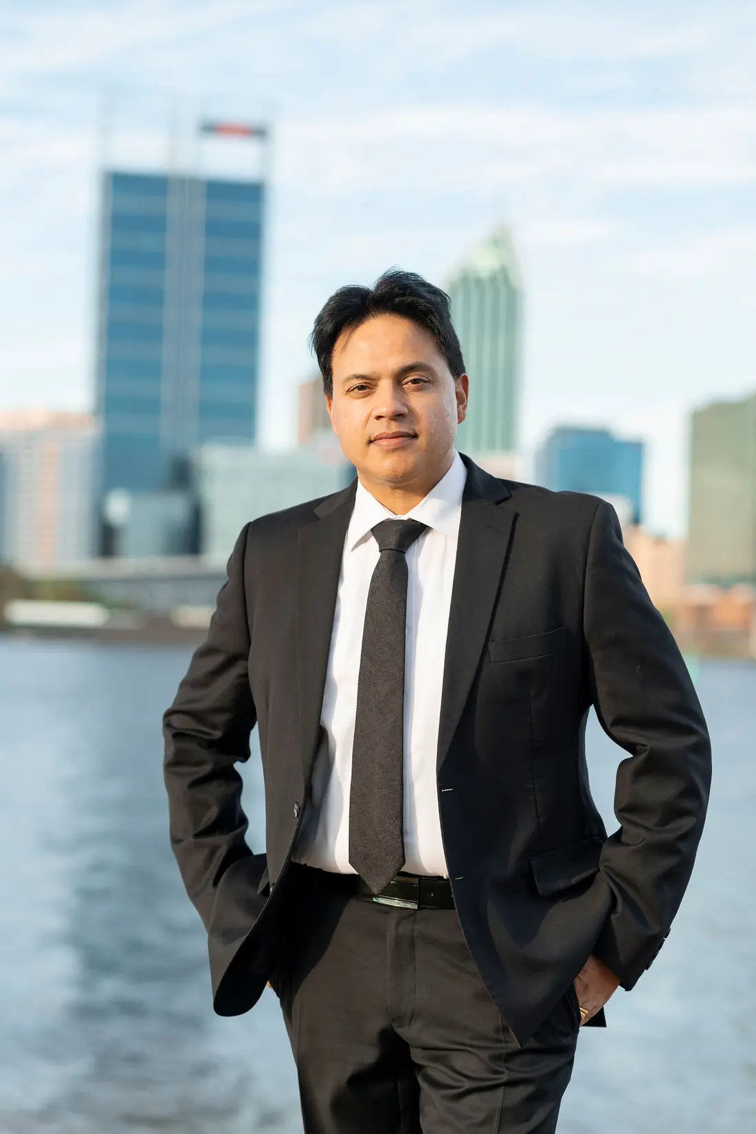 Davinder Bhogal in a dark suit on the South Perth foreshore, with the Perth city skyline behind him
